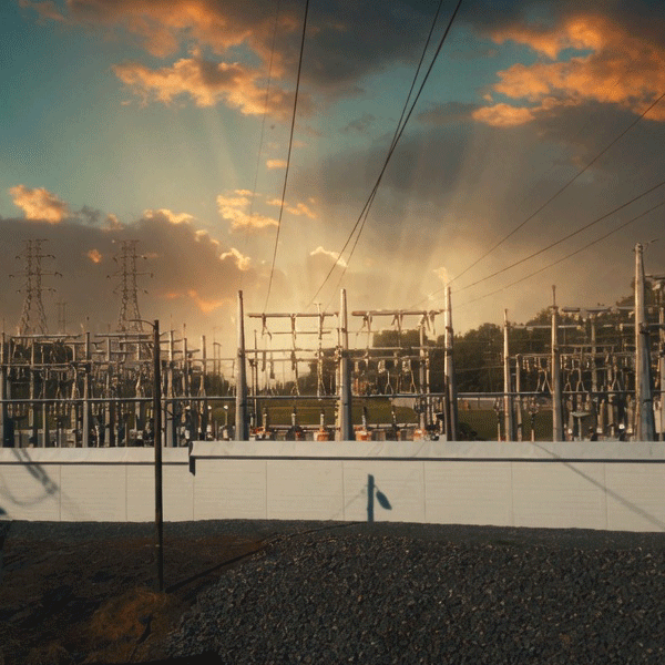 A large electrical substation and transmission yard at sunset, featuring rows of high-voltage equipment, transformers, and steel lattice towers silhouetted against a dramatic sky filled with golden sun rays breaking through dark clouds. In the foreground, a clean white solid barrier wall runs horizontally across the scene, providing visual screening and separation from the critical infrastructure behind it. Gravel ground and a partial rooftop are visible at the bottom of the frame.