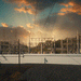 A large electrical substation and transmission yard at sunset, featuring rows of high-voltage equipment, transformers, and steel lattice towers silhouetted against a dramatic sky filled with golden sun rays breaking through dark clouds. In the foreground, a clean white solid barrier wall runs horizontally across the scene, providing visual screening and separation from the critical infrastructure behind it. Gravel ground and a partial rooftop are visible at the bottom of the frame.