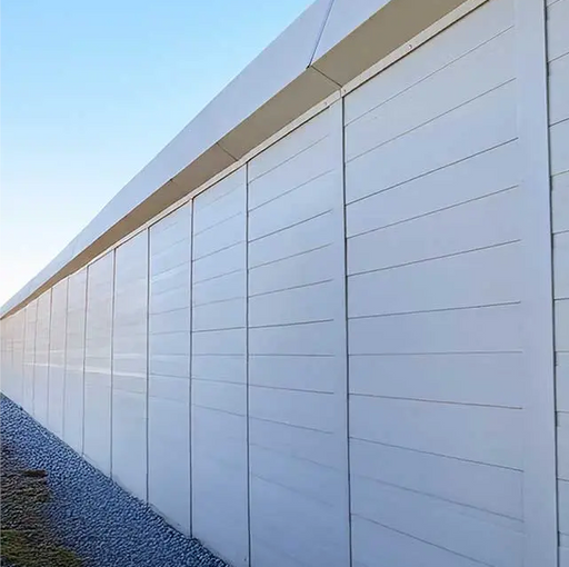A long, modern white security and privacy barrier wall with horizontal paneling stretches into the distance along a gravel path. The clean, solid panels are framed with vertical metal supports and topped by a continuous cap. Overhead power lines are visible against a clear blue sky, with sparse vegetation growing at the base of the wall.